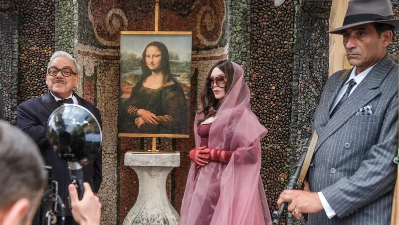 Na fotografii je slávnostná udalosť alebo rekonštrukcia scény s obrazom Mony Lisy na stojane, pred vykladanou stenou z kamienkov a mozaiky. Okolo obrazu stoja ľudia: jeden muž v obleku s motýlikom, žena v ružových šatách s červenými rukavicami a závojom, a ďalší muž v pruhovanom obleku a klobúku drží repliku samopalu. Atmosféra pôsobí ako z inscenácie historickej krádeže alebo návratu známeho obrazu, pričom v popredí je fotograf s vintage fotoaparátom