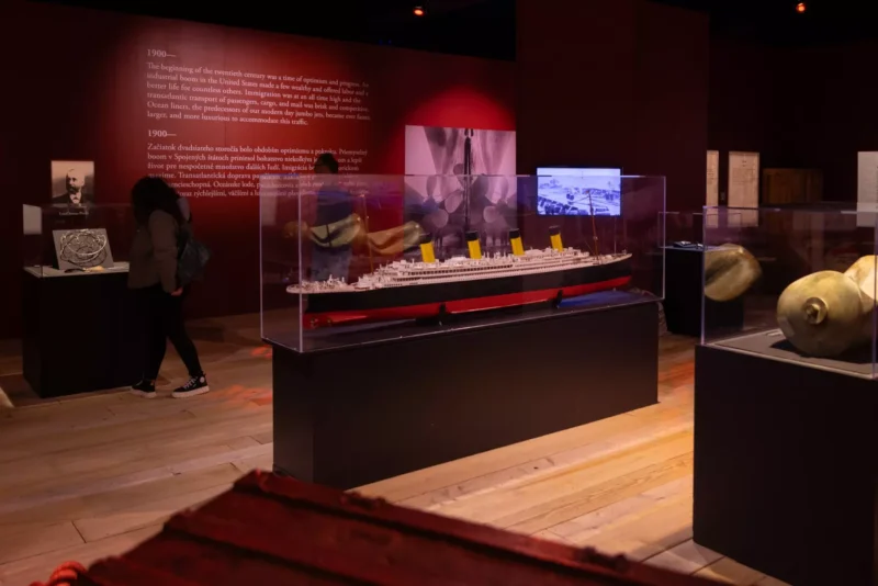 Model lode RMS Titanic zaliaty v skle s ikonickými žltými lievikmi. Pozadie obsahuje historický text a obrázky, ktoré vytvárajú nostalgickú atmosféru.