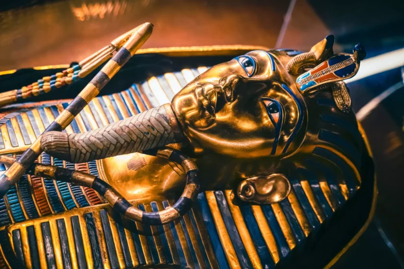 Fotografia detailne zobrazuje zlatú staroegyptskú pohrebnú masku faraóna, zdobenú tyrkysovými a modrými detailmi. Na hrudi masky sú skrížené žezlá typické pre egyptských panovníkov. Farby a lesk zlata spolu s precíznymi ornamentami podčiarkujú umeleckú hodnotu artefaktu.