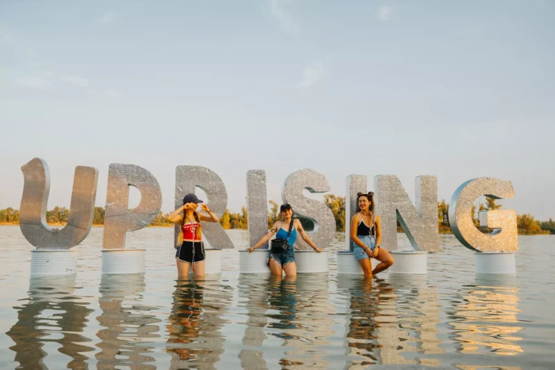 Na slnečnej letnej fotografii stoja tri dievčatá v plavkách a letnom oblečení vo vode pred veľkými písmenami „UPRISING“, ktoré sú umiestnené na podstavcoch v jazere. V pozadí je vidieť zelené stromy, jasnú oblohu a festivalovú atmosféru šťastného dňa pri vode. Scéna pôsobí zábavne, sviežo a symbolizuje atmosféru Uprising Festivalu.