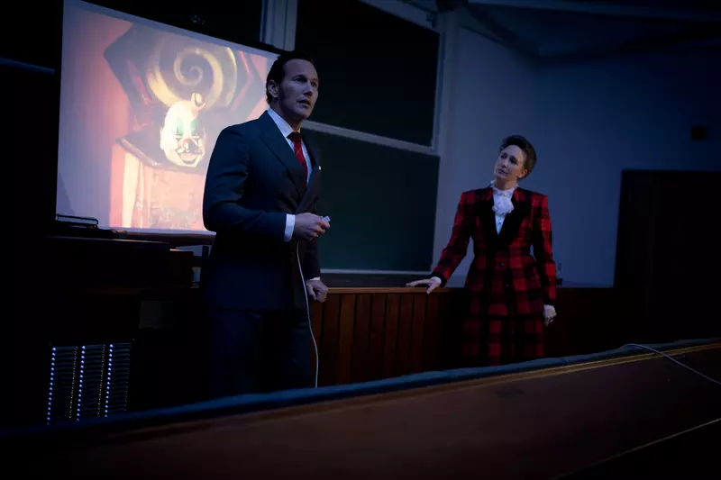 Muž v obleku prednáša v slabo osvetlenej miestnosti, na plátne sa premieta strašidelný klaun. Žena v kockovanom obleku zamyslene pozoruje.