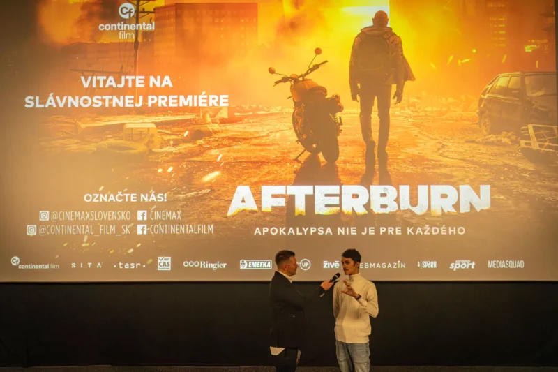 Dvaja ľudia stoja pred veľkým filmovým plátnom, na ktorom sa premieta film „Afterburn“. Scéna zobrazuje postapokalyptickú krajinu s motorkou, čo vytvára dramatickú a intenzívnu atmosféru.