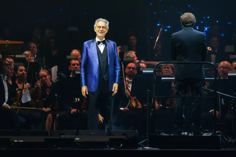 Na fotografii je elegantne oblečený sólový spevák v trblietavom modrom saku, stojaci pred veľkým symfonickým orchestrom. Dirigent sa nachádza na pódiu chrbtom k obecenstvu a vedie hudobníkov s klasickými nástrojmi. Scéna pôsobí slávnostne, profesionálne a vyzdvihuje atmosféru významného koncertu klasickej hudby.