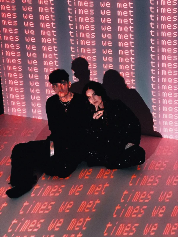 Na fotografii s moderným umeleckým nádychom sedia dvaja ľudia na podlahe, obklopení projekciou svetloružového nápisu „times we met“, ktorý je opakovane premietaný na steny aj podlahu. Oblečenie pôsobí tmavo a štýlovo, scéna je kreatívna, introspektívna a evokuje digitálnu či poetickú atmosféru spomienok.