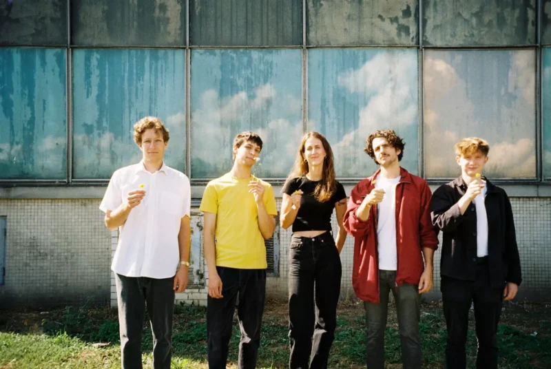 Päť ľudí stojí v rade so zmrzlinami a usmieva sa pod čiastočne zamračenou oblohou. Sú opretí o textúrovanú sklenenú stenu, čo vyžaruje uvoľnenú a radostnú náladu.