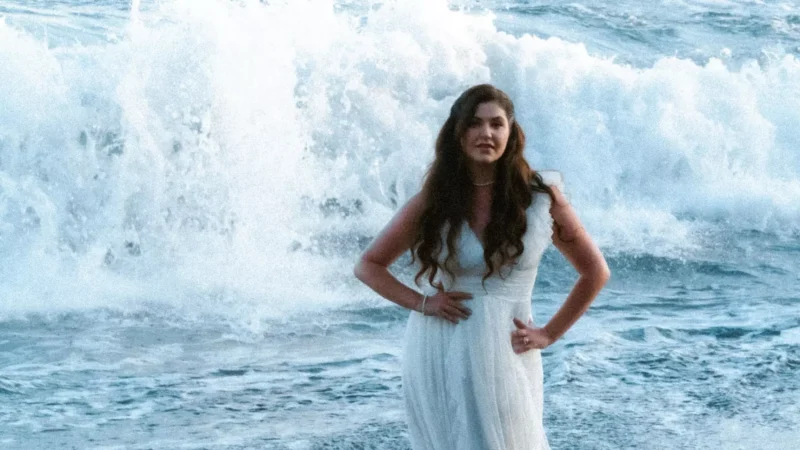 Na fotografii stojí žena v dlhých vlnitých vlasoch a bielych šatách pred dramatickou kulisou morských vĺn práve pri brehu. Dynamický záber zachytáva silu vody a kontrastuje jemnosť oblečenia s energiou mora, pričom pôsobí sviežo, snovo a romanticky.