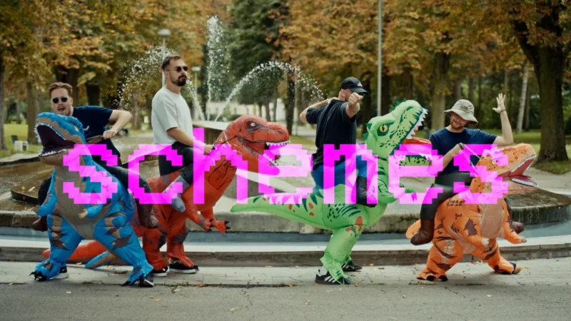 Päť ľudí v nafukovacích kostýmoch dinosaurov stojí pred fontánou s pixelovanými textovými „schemes“ v ružovej farbe. Scéna je hravá a humorná.
