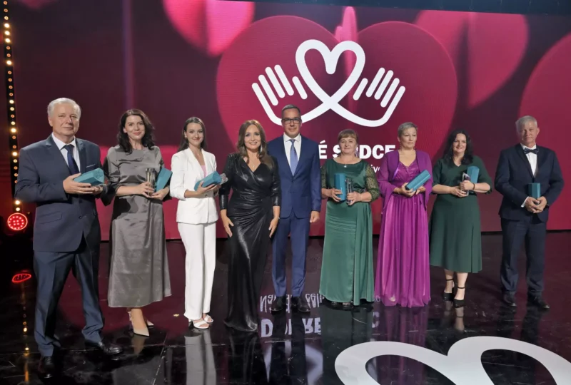 Skupina deviatich elegantne oblečených ľudí stojí na pódiu a drží modré ocenenia. Za nimi je červené pozadie s logom srdca a rúk, ktoré pripomína slávnostný ceremoniál.