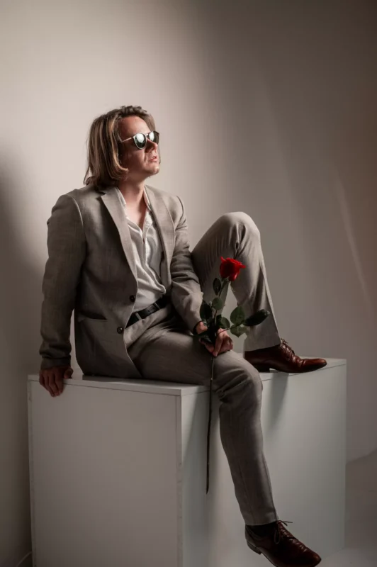Muž v béžovom obleku a slnečných okuliaroch sedí na bielej kocke v uvoľnenej póze a drží červenú ružu. Scéna je jemne osvetlená, čo vytvára štýlovú, kontemplatívnu atmosféru.