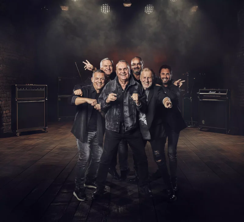 Skupina šiestich usmievavých mužov stojí v slabo osvetlenom hudobnom sále a vzrušene gestikuluje. Sú obklopení zosilňovačmi na drevenom pódiu, z ktorého vyžaruje živá a energická atmosféra.