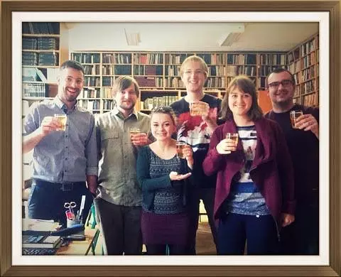 Skupina šiestich ľudí stojí s úsmevom v knižnici a drží v rukách nápoje. V pozadí sú police plné kníh, ktoré vytvárajú útulnú, slávnostnú atmosféru.