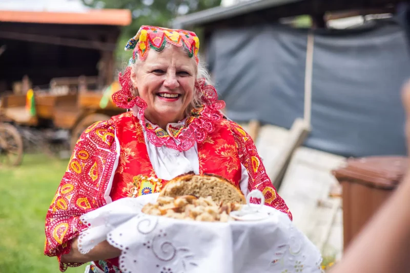 Na fotografii je žena v červenom kroji s výrazným vzorom, ktorá drží na výšivkovej utierke tanier s tradičným domácim chlebom a slaninou. V pozadí je vidiecky dvor s kočiarom a hospodárskymi stavbami, čo podčiarkuje folklórnu a slávnostnú atmosféru akcie. Scéna pôsobí pohostinne, vyjadruje tradíciu a pohodu slovenského vidieka.