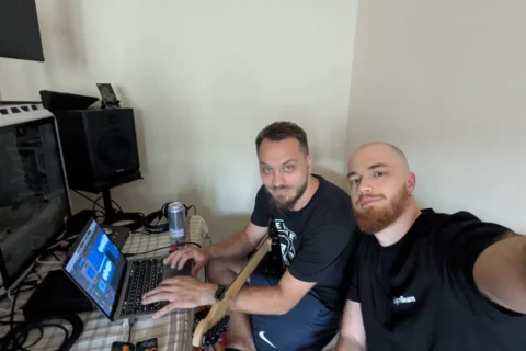 Dvaja muži sedia pri stole s notebookom a hudobným vybavením, jeden drží gitaru. Vyzerajú zaujato a uvoľnene v domácom štúdiu.