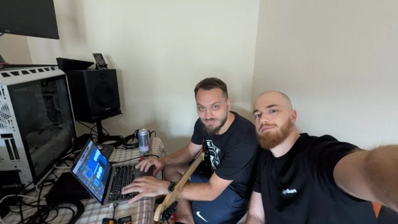 Dvaja muži sedia pri stole s notebookom a hudobným vybavením, jeden drží gitaru. Vyzerajú zaujato a uvoľnene v domácom štúdiu.
