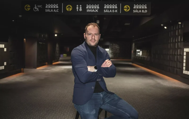 Muž v tmavom saku a džínsoch sedí v slabo osvetlenej, modernej hale kina. Nad hlavou sú tabule s nápismi, ktoré ukazujú smer rôznych kín. Atmosféra je pokojná a sofistikovaná.