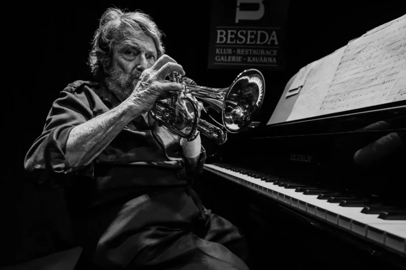 Čiernobiela fotografia zachytáva hudobníka sediaceho pred klavírom Petrof so skladbou na pulte, ktorý hrá na trúbku alebo krídlovku. V pozadí je tabuľa klubu Beseda. Kompozícia pôsobí intímne, sústredene a evokuje atmosféru koncertu v malom hudobnom klube.