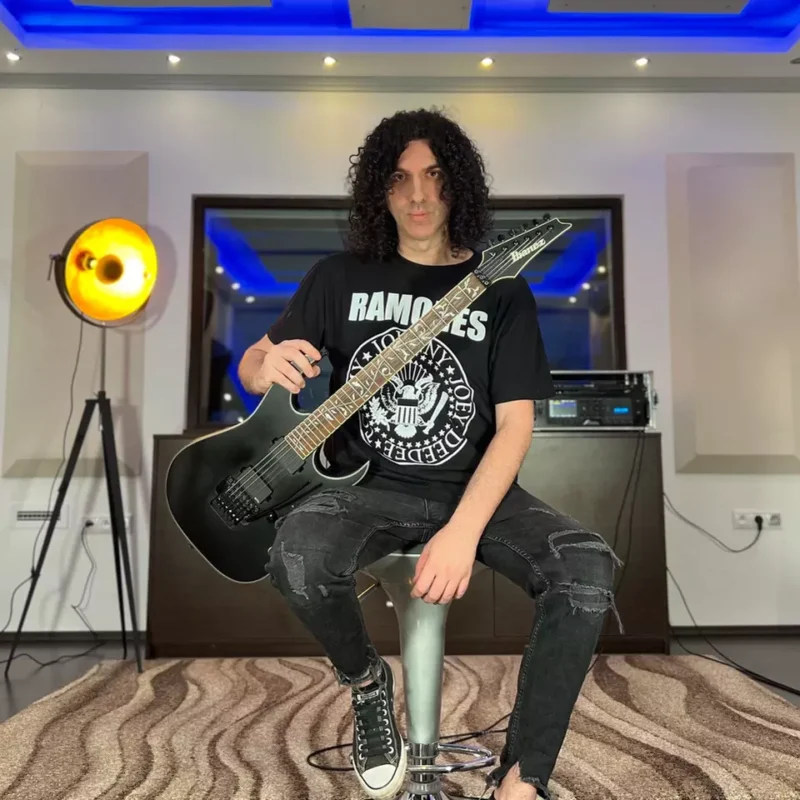 Muž s kučeravými vlasmi sedí na stoličke v modernej miestnosti a drží čiernu elektrickú gitaru. V tričku s nápisom „Ramones“ pôsobí uvoľnenou rockovou atmosférou.