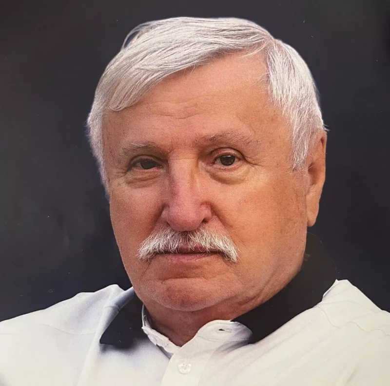 Starší muž s bielymi fúzikmi a vlasmi, oblečený v bielej košeli s čiernym golierom. Má vážny výraz na tmavom pozadí.