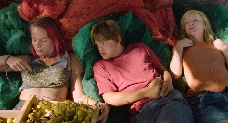 Traja ľudia ležia vonku na farebných rybárskych sieťach, dve ženy a jeden mladík. Scéna pôsobí uvoľnene, s hroznom v drevenej debne neďaleko, čo vyvoláva pocit pokoja a priateľstva.