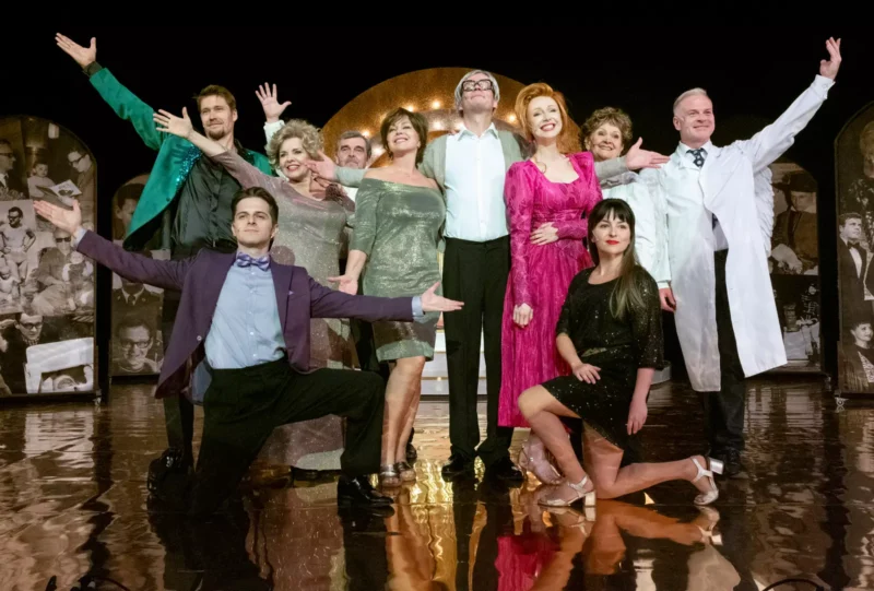 Rôznorodá skupina deviatich jednotlivcov v žiarivých, elegantných outfitoch pózuje expresívne na pódiu a vyžaruje radosť a oslavu. V pozadí sú historické fotografie.