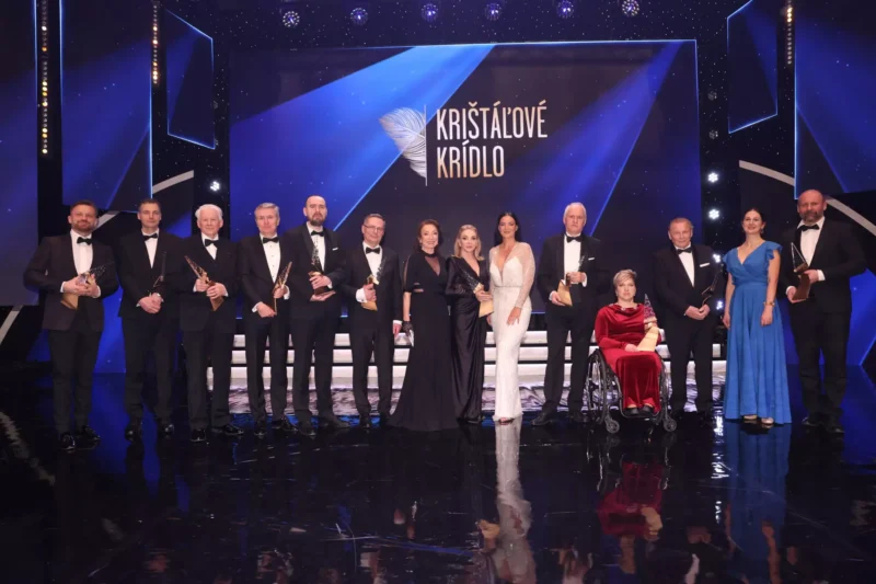 Skupina elegantne oblečených mužov a žien pózuje na pódiu a drží ocenenia na podujatí „Krištáľové krídlo“. Pozadie je modré s trblietavými svetlami.