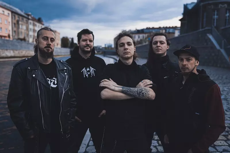 Päť mužov sebavedomo stojí na dláždenom brehu rieky. Sú oblečení v tmavom, ležérnom oblečení, čo svedčí o vážnej a intenzívnej nálade. V pozadí je zamračená obloha a mestské budovy.