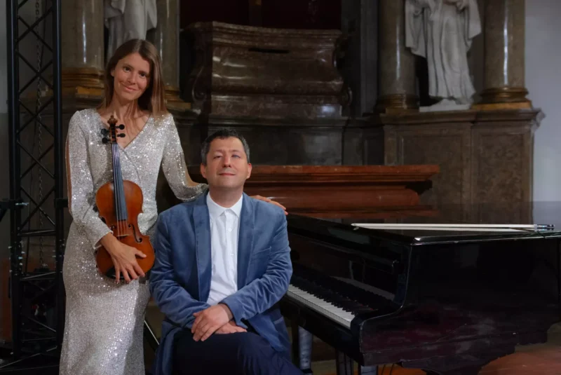 Žena v trblietavých šatách drží husle a stojí vedľa sediaceho muža v modrom saku pri klavíri v elegantnej historickej miestnosti.