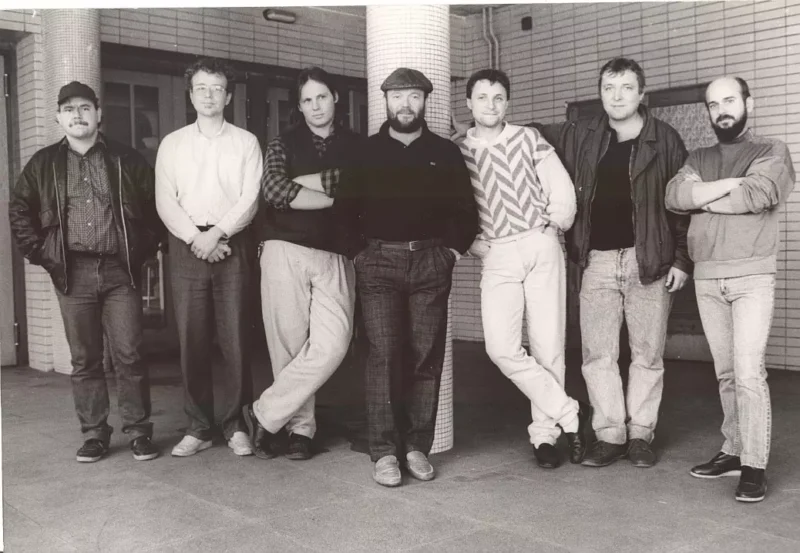 Čiernobiela fotografia siedmich mužov ležérne stojacich v rade v interiéri. Majú na sebe ležérne oblečenie s neutrálnymi výrazmi tváre, čo vytvára uvoľnenú atmosféru.