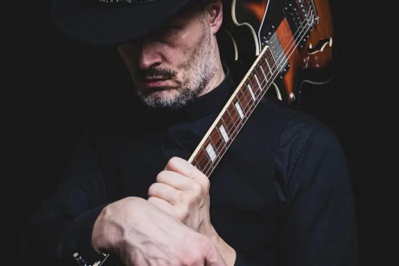 Muž v čiernom kovbojskom klobúku a košeli si drží gitaru opretú o plece. Na tmavom pozadí hľadí dole s vážnym výrazom.