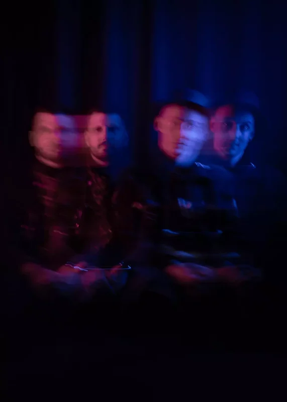 Rozmazaný obraz štyroch osôb sediacich na tmavom pozadí, osvetlený modrými a červenými odtieňmi, ktorý vyvoláva tajomnú, snovú náladu.