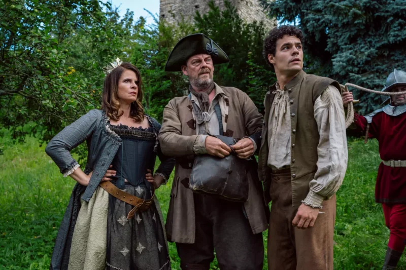 Fotografia zobrazuje skupinu ľudí v historických kostýmoch, stojacich na tráve pred hradnou stenou a hustou zeleňou. Jeden muž má trojrohý klobúk a tašku, žena je v korzete so sukňou a opaskom, ďalší muž v košeli a veste; v pozadí je viditeľný strážca v brnení s halapartňou. Scéna pôsobí ako výjav z dobového filmu alebo divadelnej inscenácie zasadený do stredovekých či ranonovovekých čias.