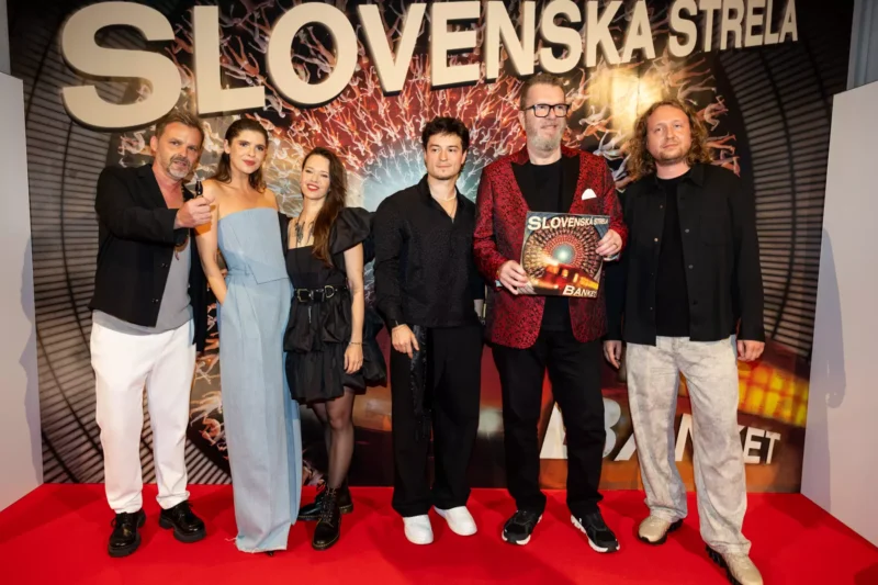 Šesť ľudí stojí na červenom koberci a usmieva sa na podujatí. Muž v červenej bunde drží nápis „Slovenská strela“. Za ním je viditeľné veľké logo.