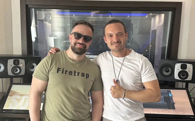 Dvaja muži stoja a usmievajú sa v nahrávacom štúdiu. Jeden má slnečné okuliare a tričko s nápisom „Firetrap“, druhý má na sebe biele oblečenie s palcom hore. Za nimi sú reproduktory a ozvučnica.