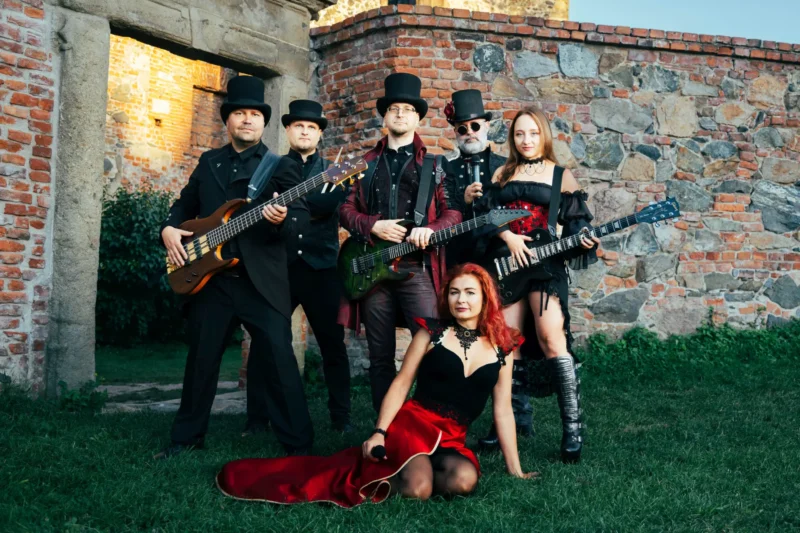 Kapela v steampunk oblečení pózuje s nástrojmi na pozadí kamennej steny. Sediaca žena v červených šatách drží mikrofón a vyžaruje sebavedomie.