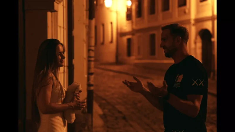 Muž a žena sa vrúcne rozprávajú na dláždených uliciach v noci, osvetlených jemnými zlatými pouličnými lampami. Scéna vyvoláva priateľskú a intímnu atmosféru.