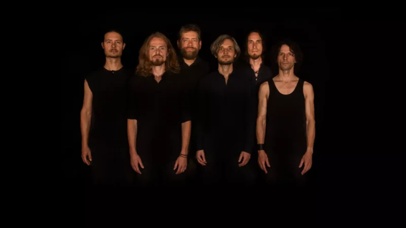 Šesťčlenná skupina stojí v dvoch radoch, každý je oblečený v jednoduchom čiernom oblečení na tmavom pozadí. Kompozícia pôsobí dramaticky a sústredene, zvýrazňuje kolektívnu energiu a jednotu členov, zároveň je vizuálne minimalistická a silná.