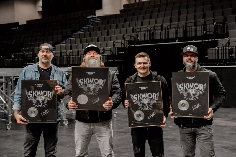 Štyria muži stoja v prázdnom koncertnom sále, každý drží zarámovanú plaketu s nápisom „Škwor“ a CD. Vyzerajú hrdo a veselo.