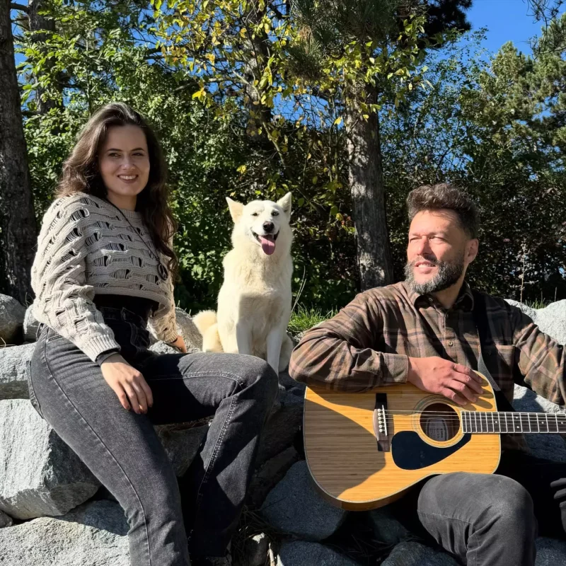 Muž s gitarou, žena a biely pes sedia na skalách v slnkom zaliatom lese. Scéna je uvoľnená a radostná pod jasnou modrou oblohou.
