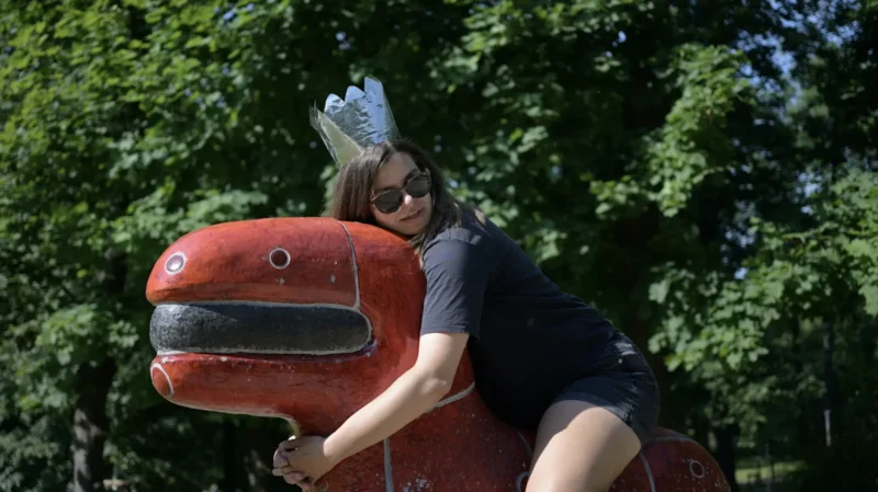 Osoba so slnečnými okuliarmi a striebornou korunou vonku objíma veľkú červenú sochu rozmarného stvorenia. Nálada je hravá, v pozadí svieže zelené stromy.