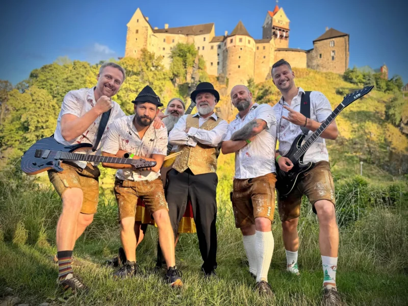 Radostná skupina šiestich mužov v tradičnej póze z kožených nohavíc s gitarami pred hradom na kopci pod jasnou modrou oblohou, vyžarujúca kamarátstvo.