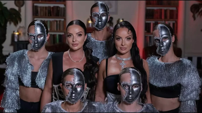 Dve ženy v elegantných čiernych šatách a strieborných šperkoch pózujú so štyrmi ďalšími v strieborných maskách a strapcových topoch v miestnosti s knižnicou. Nálada je očarujúca a tajomná.