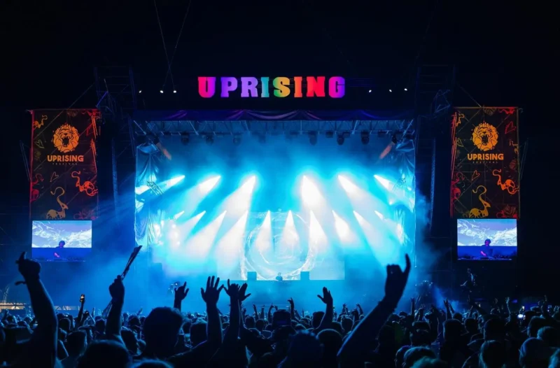 Živá koncertná scéna s farebným transparentom „Uprising“. Jasnomodré svetlá pódia osvetľujú veľký dav so zdvihnutými rukami a vytvárajú energickú atmosféru.