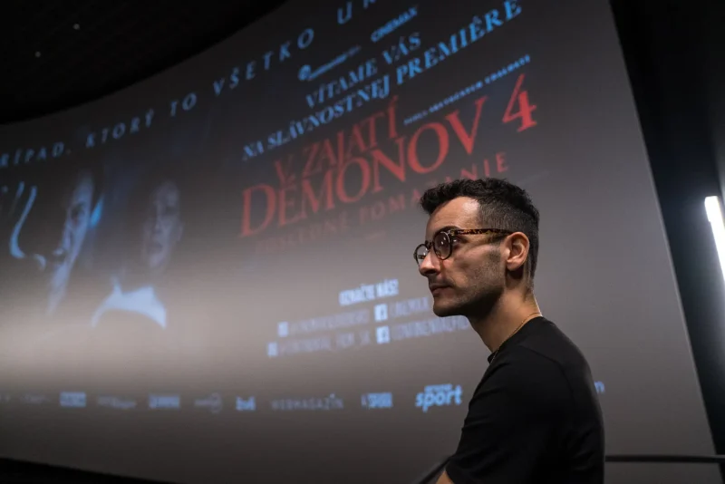 Muž s okuliarmi stojí v tmavom kine pred veľkou obrazovkou, na ktorej je zobrazený filmový plagát k filmu „V Zajatí Démonov 4“, čím vytvára napínavú atmosféru.