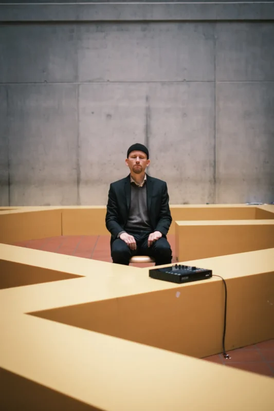 Osoba sedí v kontemplatívnej póze na stoličke v geometrickej žltej lavici opretej o betónovú stenu. Na lavici je malý zvukový mixážny pult.