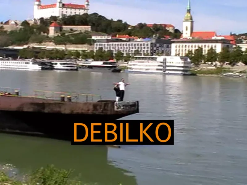 Pár napodobňuje pózu Titanicu na móle pri rieke. V pozadí sú lode a historická panoráma mesta. Prekrytie textom znie „DEBILKO“.