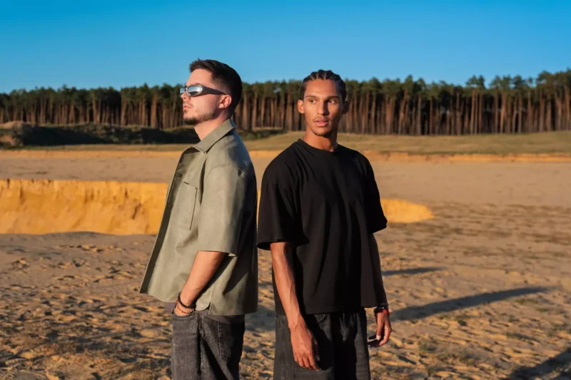 Dvaja muži stoja chrbtom k sebe v piesočnatej krajine pod jasnou modrou oblohou. Jeden má slnečné okuliare a zelenú košeľu, druhý čiernu košeľu. V pozadí les.