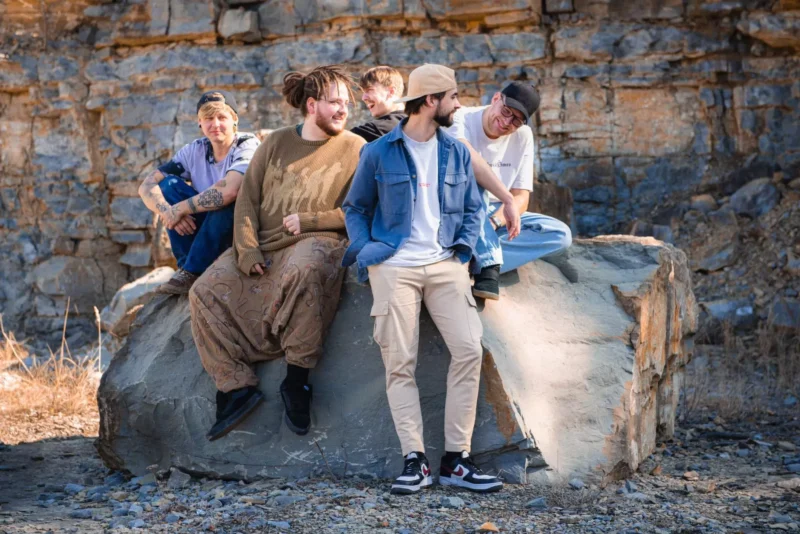 Päť priateľov ležérne sedí na veľkom skale v kameňolome. Sú oblečení v uvoľnenom, zemitom oblečení a zdá sa, že sa smejú a rozprávajú. Prostredie je drsné a vytvára bezstarostnú a hravú atmosféru.