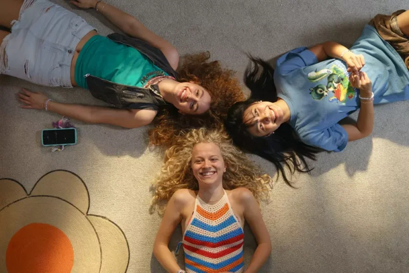 Tri ženy ležia v kruhovom útvare na koberci a radostne sa usmievajú. Jedna má na sebe háčkovaný top, druhá tričko s potlačou a neďaleko leží telefón.