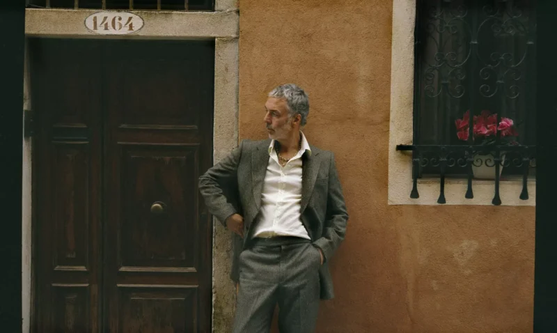 Muž v sivom obleku stojí ležérne s jednou rukou vo vrecku, opretý o hnedé drevené dvere. Prostredie tvorí rustikálna oranžová stena s oknom ozdobeným červenými kvetmi, ktorá vyvoláva uvoľnenú a nostalgickú náladu.