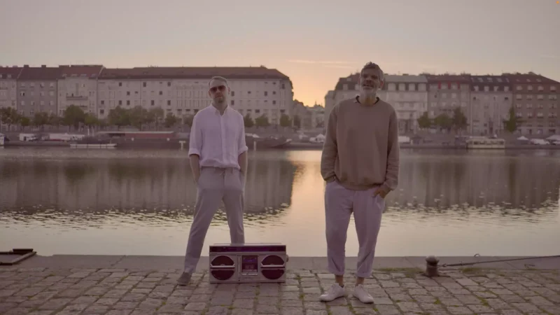 Dvaja muži stoja na brehu rieky pri západe slnka, v pozadí pastelovo sfarbené budovy. Jeden má na sebe bielu košeľu a druhý béžový sveter. Medzi nimi je na zemi boombox, ktorý vytvára pokojnú a nostalgickú atmosféru.